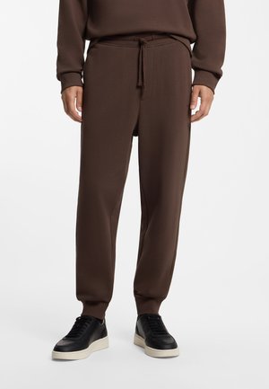 Hombre que lleva pantalones jogger marrones con puños elásticos y cintura con cordón, combinados con zapatillas negras de corte bajo con suelas blancas.