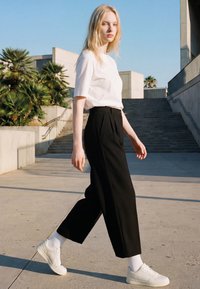 Young woman walks outdoors near stairs, wearing white t-shirt, black wide-leg pants, white socks, and white sneakers in daylight.
