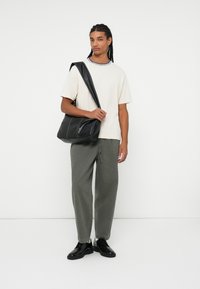 Sort læder skuldertaske med en glat overflade, pyntet med subtile syninger, kombineret med en beige t-shirt og olivengrønne bukser.