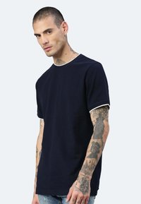 T-shirt in cotone blu navy con maniche corte e colletto a costine bianche. Il modello ha tatuaggi visibili sulle braccia e indossa jeans blu sbiaditi.