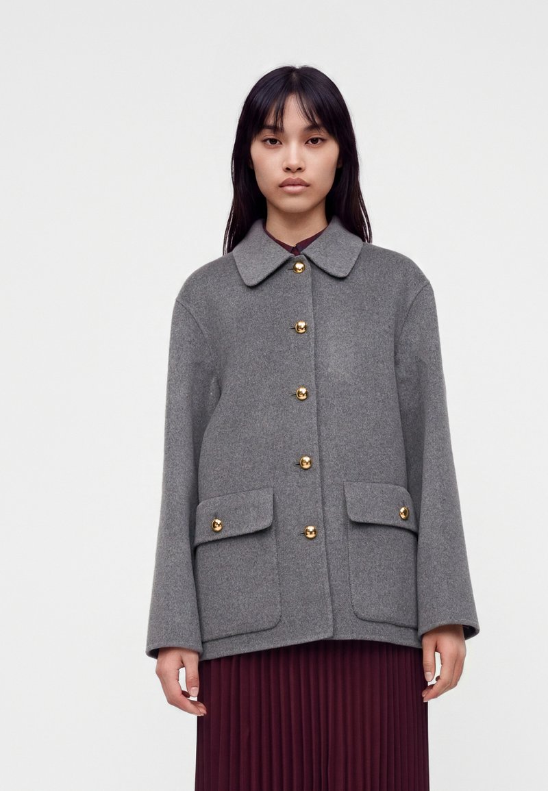 Femme portant un manteau en laine gris boutonné avec des boutons dorés et des poches avant sur une jupe plissée bordeaux, debout devant un fond uni.