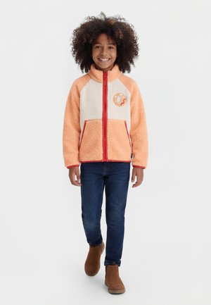 Barn med krøllet hår går fremad iført orange og creme fleecejakke med tegneserie-patch, mørke jeans og brune støvler.