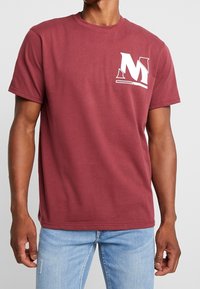 Bordeaux-farvet bomulds T-shirt med korte ærmer, rund hals og en hvid "M" logo på venstre bryst. Stoffet ser blødt og let tekstureret ud.