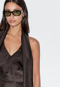 Top nero senza maniche con una trama stropicciata e scollatura a V. Il modello indossa occhiali da sole rettangolari verde scuro. Stile minimalista, sfondo neutro.