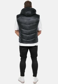Gilet puffer in pelle nera con collo alto, caratterizzato da un motivo trapuntato e un orlo curvo, indossato sopra una camicia bianca e jeans neri.