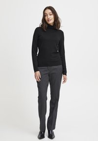 Maglione a collo alto nero abbinato a pantaloni grigi slim-fit con texture e stivaletti neri. L'outfit ha una silhouette aderente.