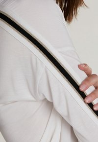 Camicia bianca a maniche lunghe con un accento a strisce nere e dorate metalliche lungo la cucitura della spalla. Tessuto liscio e leggero.