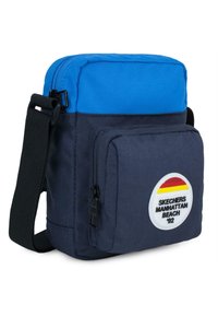 Bandolera azul y oscura de tela, con un bolsillo frontal con cremallera y un parche circular con un diseño retro.