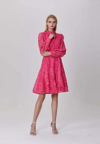 Femme blonde debout, portant une robe en dentelle rose à manches longues avec des épaules bouffantes et des talons blancs à bride de cheville sur un fond uni.