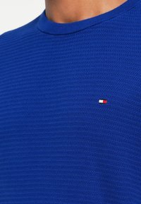 Kék, textúrázott pulóver bordázott mintával és kerek nyakkal, a mellrészen egy kis piros, fehér és kék logóval.