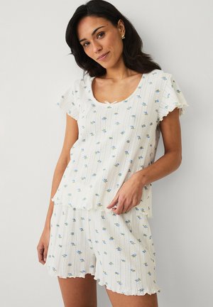 Kvinde iført hvid kortærmet top og matchende shorts med lille blåt blomsterprint, stående mod en ensartet lys baggrund.