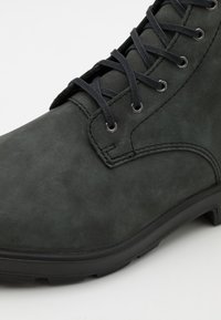 Stivale alla caviglia in camoscio verde scuro con lacci neri, dettagli cuciti e suola in gomma. Presenta occhielli metallici e una texture liscia.