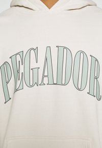 Felpa con cappuccio beige chiaro con scritta in verde audace e curva "PEGADOR". Tessuto morbido con cappuccio con coulisse, senza motivi visibili.