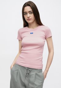 Różowy dopasowany t-shirt z prążkowanym wzorem, krótkimi rękawami i niebieską naszywką z logo "HUGO" na przodzie, w zestawie z szarymi spodniami na sznurku.