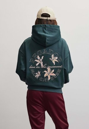 Persona che indossa una felpa con cappuccio verde scuro con motivo floreale e testo "PEGADOR live in the moment" vista da dietro, cappello beige e pantaloni color granata.