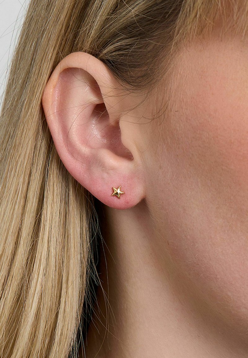 Gros plan sur l'oreille d'une femme aux cheveux blonds lisses, portant une petite boucle d'oreille en forme d'étoile en or sur le lobe de l'oreille.