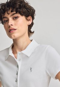 Vit polo skjorta med en strukturerad yta, utrustad med en knappad krage och en liten broderad design på bröstet i en kontrasterande färg.