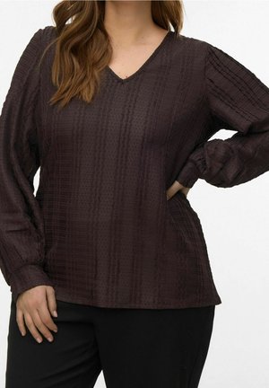 Kvinde iført en mørkebrunt tekstureret langærmet V-hals bluse med løse ærmer og sorte bukser, hånd på hoften og afslappet holdning.