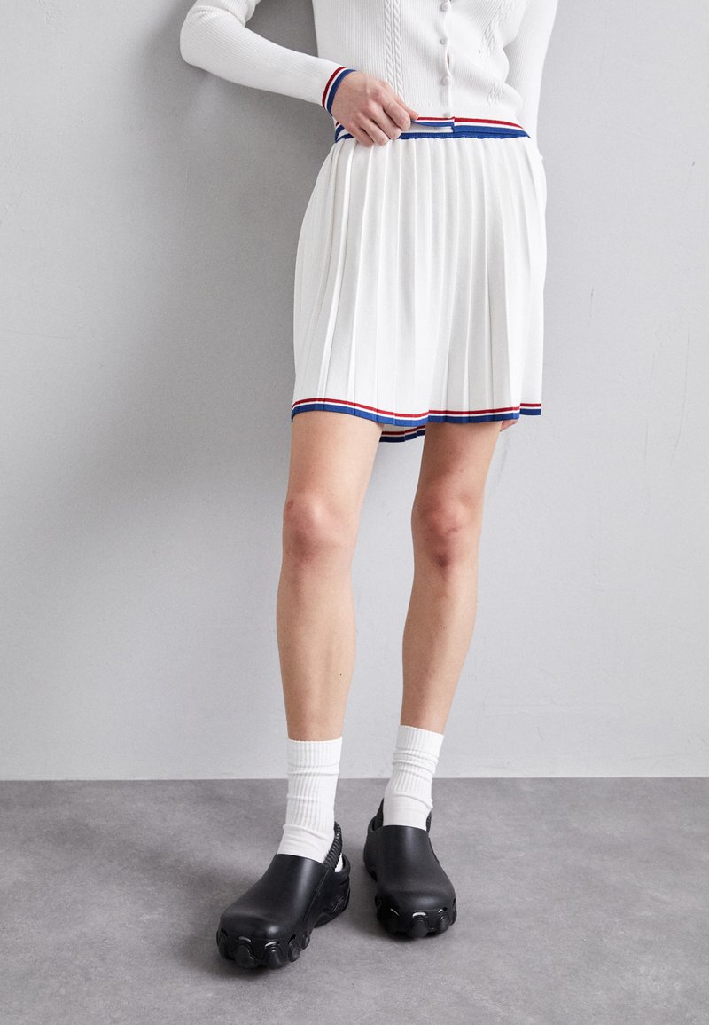 Jupe plissée blanche avec un ourlet rouge et bleu, dotée d'une taille contrastante. Assortie avec des sabots noirs et des chaussettes blanches côtelées.