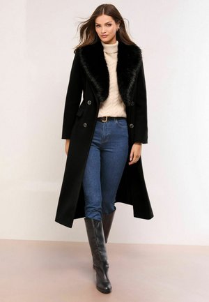 Black wool coat with a faux fur collar, double-breasted design, paired with a cable-knit cream sweater and blue jeans, finished with tall black boots.