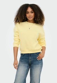 Femme aux cheveux bouclés portant un sweat-shirt jaune clair et un jean bleu, debout avec une main sur la hanche et l'autre détendue.