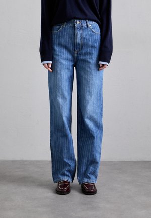Persoon draagt hoge taille blauwe wijde pijpen jeans met krijtstrepen, een zwarte trui en donker bordeauxrode loafers met studs, staand op een grijze vloer.