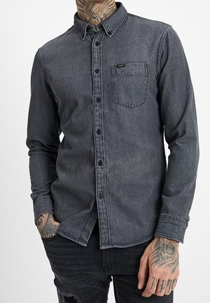 Homme portant une chemise en jean gris foncé à boutons, manches longues et poche poitrine, associée à un jean noir déchiré.