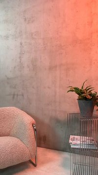 Zaokrąglony beżowy fotel boucle obok minimalistycznego metalowego stołu z drutu, na którym znajduje się doniczkowa roślina i stosy magazynów, na tle betonowej ściany.