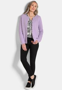 Vrouw met grijs haar, gekleed in een lila knoptrui, zwart gebloemd blouse, zwarte skinny broek en zwarte sneakers, staande voor een witte achtergrond.