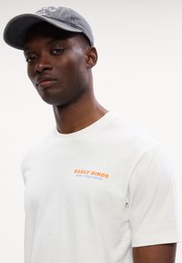 Λευκό βαμβακερό T-shirt με στρογγυλή λαιμόκοψη, που φέρει πορτοκαλί και μπλε κείμενο "EARLY BIRDS MARCO POLO DENIM." Συνδυασμένο με μια γκρι καπέλο.