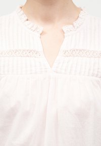 Λευκό πουκάμισο με γιακά με βολάν, κάθετες ρίγες και δαντέλα στο πάνω μέρος του στήθους, φοράει ένα άτομο που απεικονίζεται από τον λαιμό έως το μέσο του κορμού.