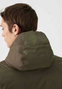 Veste rembourrée vert olive avec un col haut, dotée d'un logo en relief sur la capuche. Texture lisse avec des coutures presque invisibles.