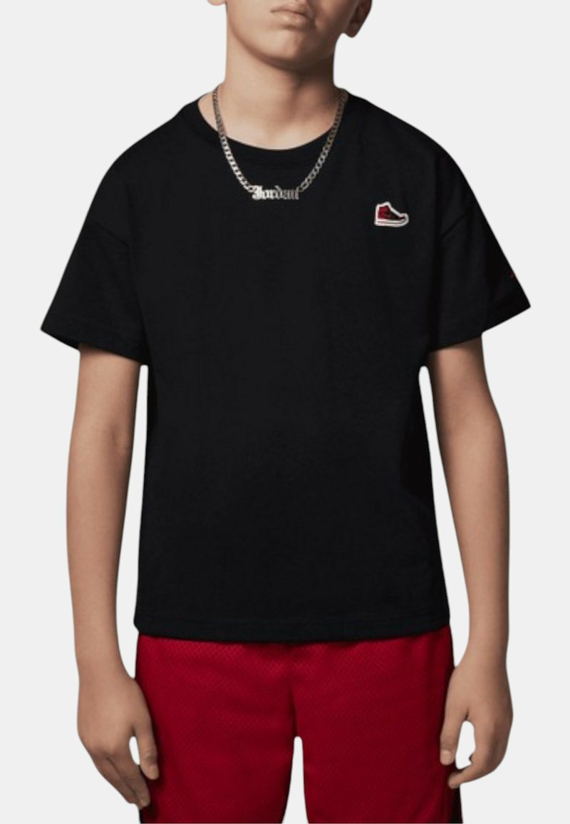 T-shirt in cotone nero con vestibilità rilassata, caratterizzata da un piccolo logo di sneaker rosso e bianco sul petto. Abbinata a pantaloni corti rossi.