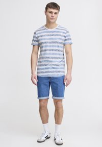 Stribet t-shirt i lys blå og hvid, parret med denimshorts. Har opkrøllede ærmer. Fodtøj inkluderer hvide sneakers med sorte accenter.