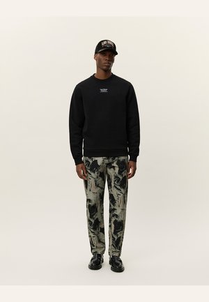 Sudadera negra con cuello redondo, combinada con pantalones holgados que presentan un patrón abstracto en verde y negro. Zapatillas negras y una gorra a juego.