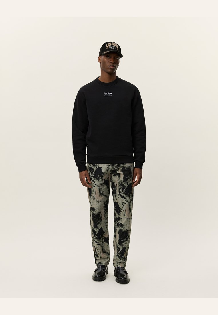 Sudadera negra con cuello redondo, combinada con pantalones holgados que presentan un patrón abstracto en verde y negro. Zapatillas negras y una gorra a juego.