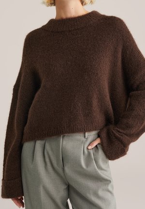 Personne portant un pull ample, marron foncé duveteux et un pantalon plissé taille haute gris clair, une main dans la poche.