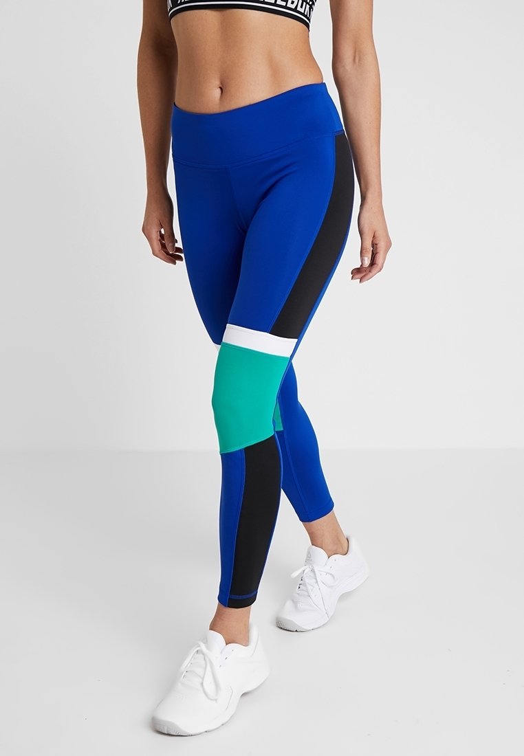 Blå träningsleggings med svarta och gröna detaljer, hög midja och figurnära design. Matchas med vita sneakers.