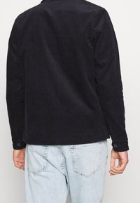 Mörk marinblå kordrock med långa ärmar, knappmanchetter och vertikal ribbtextur. Något längre bak med rund nederkant.