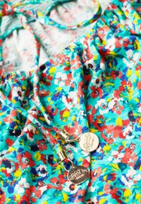 Jasny kwiatowy materiał w odcieniach turkusu, czerwieni, żółci, bieli i niebieskiego z dwiema złotymi metalowymi metkami ze znakiem marki przymocowanymi do sznurków.