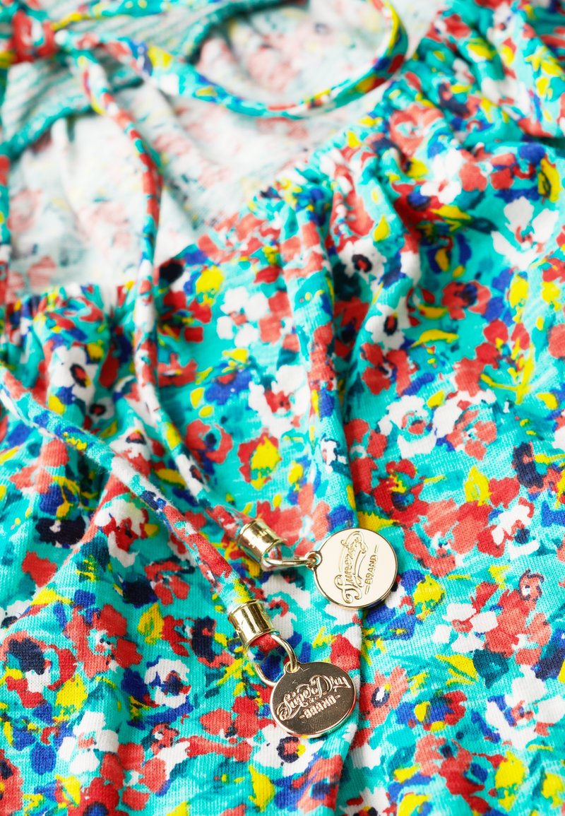 Jasny kwiatowy materiał w odcieniach turkusu, czerwieni, żółci, bieli i niebieskiego z dwiema złotymi metalowymi metkami ze znakiem marki przymocowanymi do sznurków.