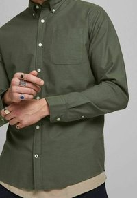 Dark green button-up shirt with a chest pocket, long sleeves, and white buttons. Worn over a light-colored shirt, hands adorned with rings.