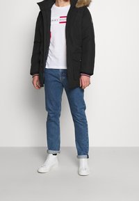 Schwarze Parkajacke mit einer mit Fell gefütterten Kapuze, Reißverschluss vorne und Taschen, getragen über einem weißen Hemd und blauen Jeans. Das Modell trägt weiße Sneakers.