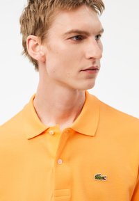 Oranssipaita, jossa on tekstuuri, kahden napin nappilista ja vihreä krokotiililogo rinnassa. Lähikuva paidan kauluksesta.
