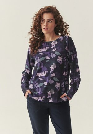 Kobieta o kręconych włosach, ubrana w długi rękaw z ciemnym kwiatowym wzorem i ciemne spodnie, stoi z rękami w kieszeniach, patrząc w prawo.