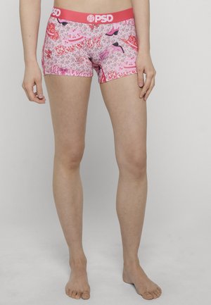 Persona con bóxers rosa PSD con patrones de corazones, zapatos y flores, de pie descalza contra un fondo claro y sencillo.