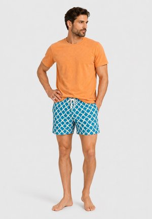 Mann steht barfuß und trägt ein oranges kurzärmeliges Shirt sowie blaue gemusterte Badeshorts mit weißem Kordelzug.
