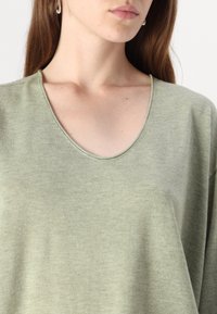 Ljusgrön, mjuk tygöverdel med bred båtringning och avslappnad passform; slät textur med subtil variation i nyansen.