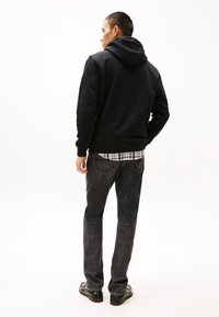 Schwarze Kapuzenjacke mit strukturierter Oberfläche, kombiniert mit dunklen Jeans. Die Jacke hat eine vordere Tasche und eine Kordelzugkapuze.