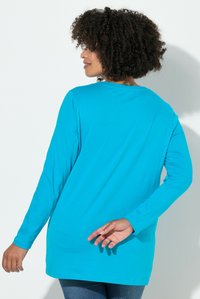 Frau mit lockigem Haar, die ein langärmeliges, leuchtend blaues Shirt und Jeans trägt, seitlich stehend mit einer Hand hinter dem Rücken.